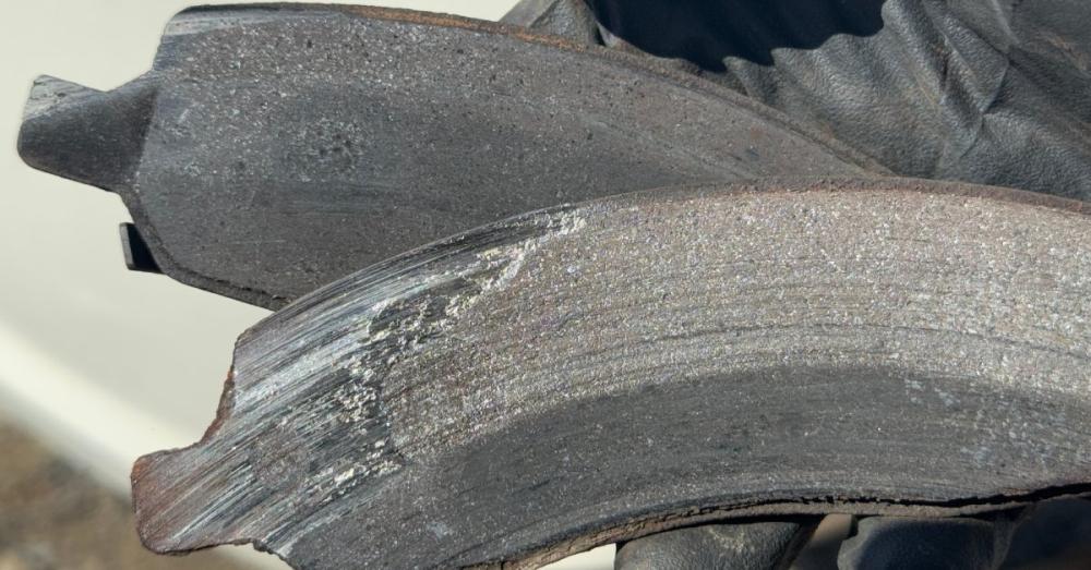 A close-up of a gloved hand holding two brake pads. The pads are separated and one is clearly more worn than the other.