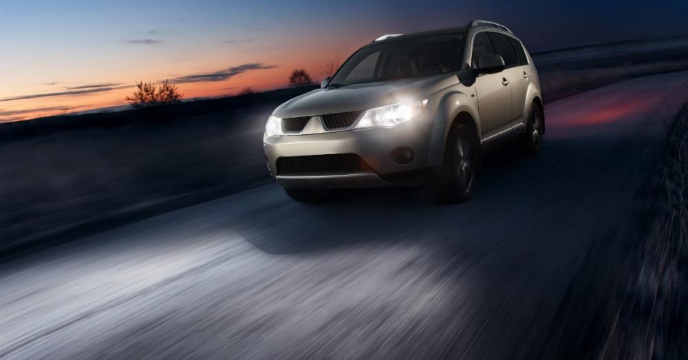 How To Replace Your Car’s Headlight Assembly A silver SUV drives on a rural road at dusk with bright headlights illuminating the pavement under a colorful sunset sky.