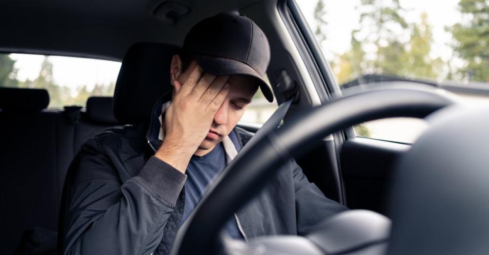 A man with a hat sitting behind the wheel of his car. He has his hand on his face and looks disappointed.