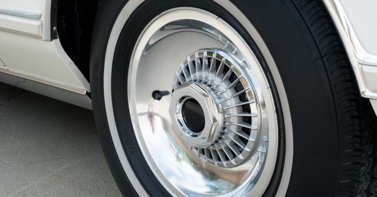 A black tire has a thick white stripe on the sidewall. The wheel has a circular center cap and closely spaced spokes.