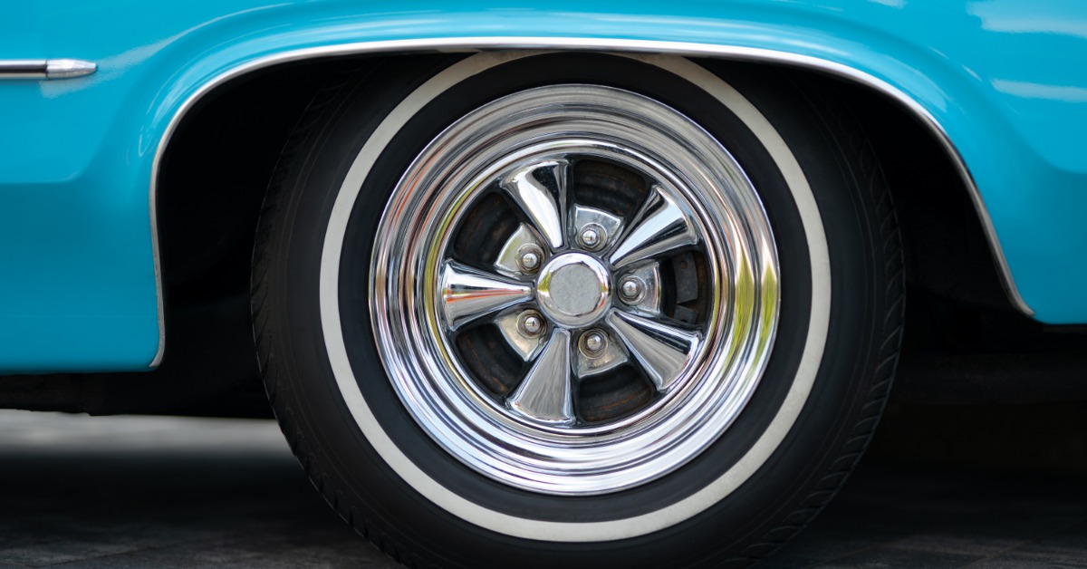 A turquoise vehicle has a white wall tire paired with a shiny five-spoke wheel. Metal trim outlines the wheel opening.