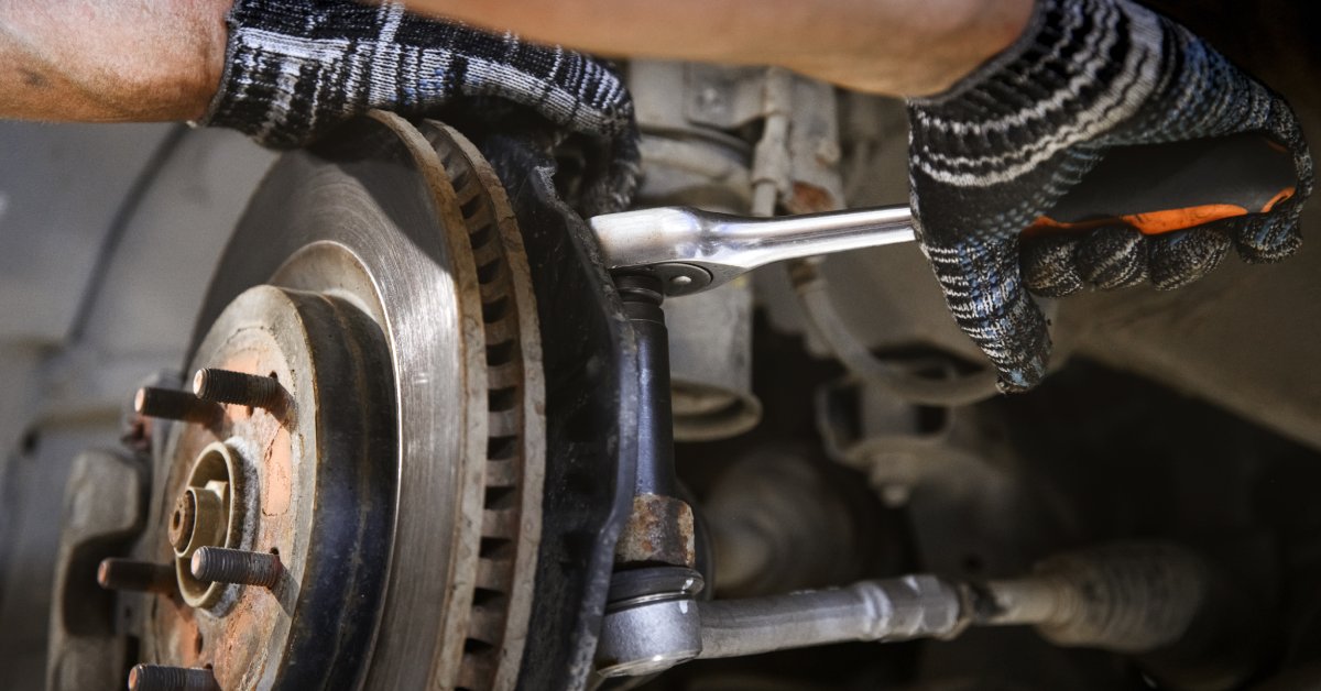 A person wearing gloves works on a car's brake system. They're using a socket wrench on the back of the rotor.