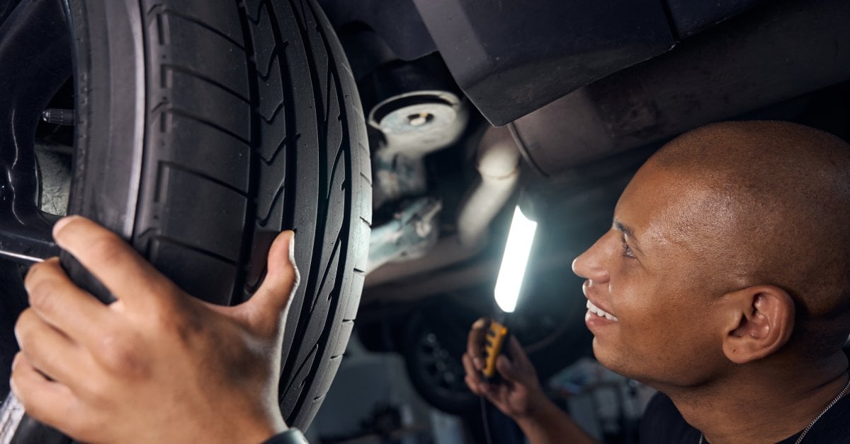 A mechanic standing under a car while holding a light in his hand. He's smiling while looking at the wheel of the car.