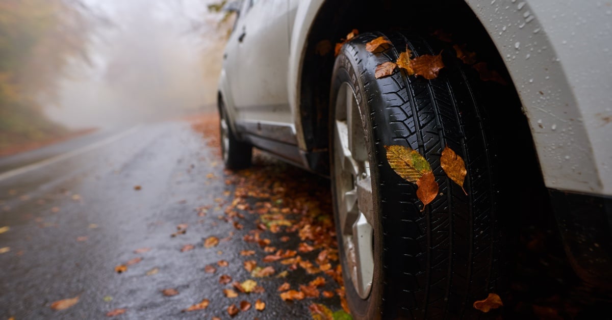 The side view of a car driving down the road. There are autumn leaves all over the ground and the tires of the vehicle.