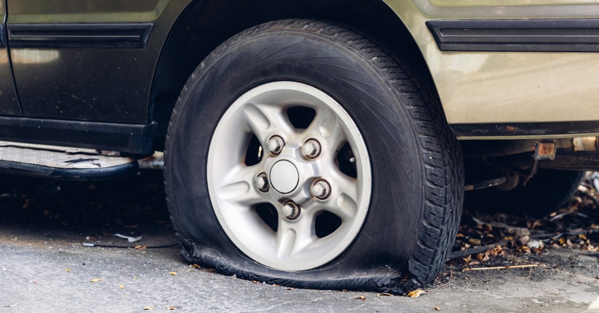 A green car on the side of the road with a flat tire. The tire is starting to fall apart due to the damage sustained.