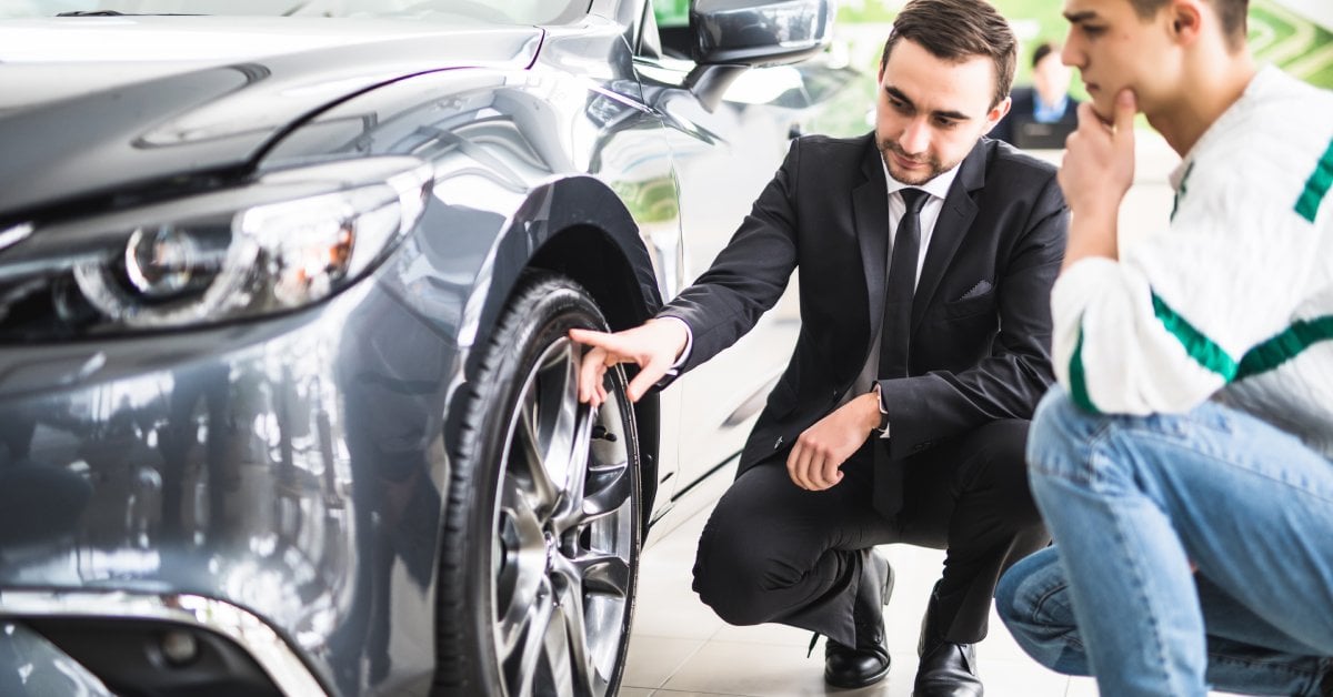 Two men, crouched down, looking at the wheel of a car. One is in a suit and the other is in casual clothes.