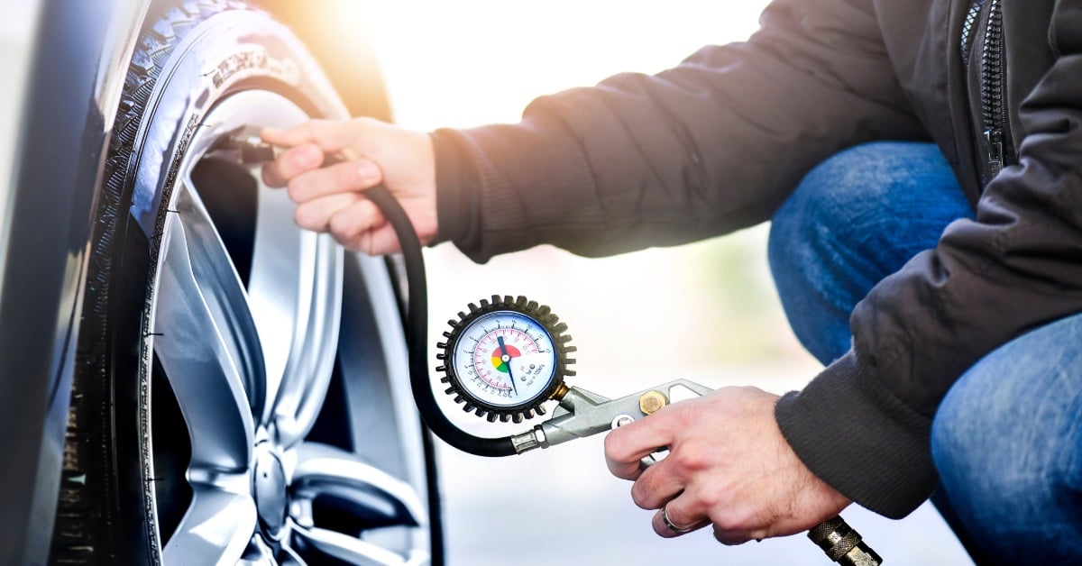 A man wearing a black puffy jacket and jeans uses a tire pressure gauge to check and inflate his vehicle’s tire.