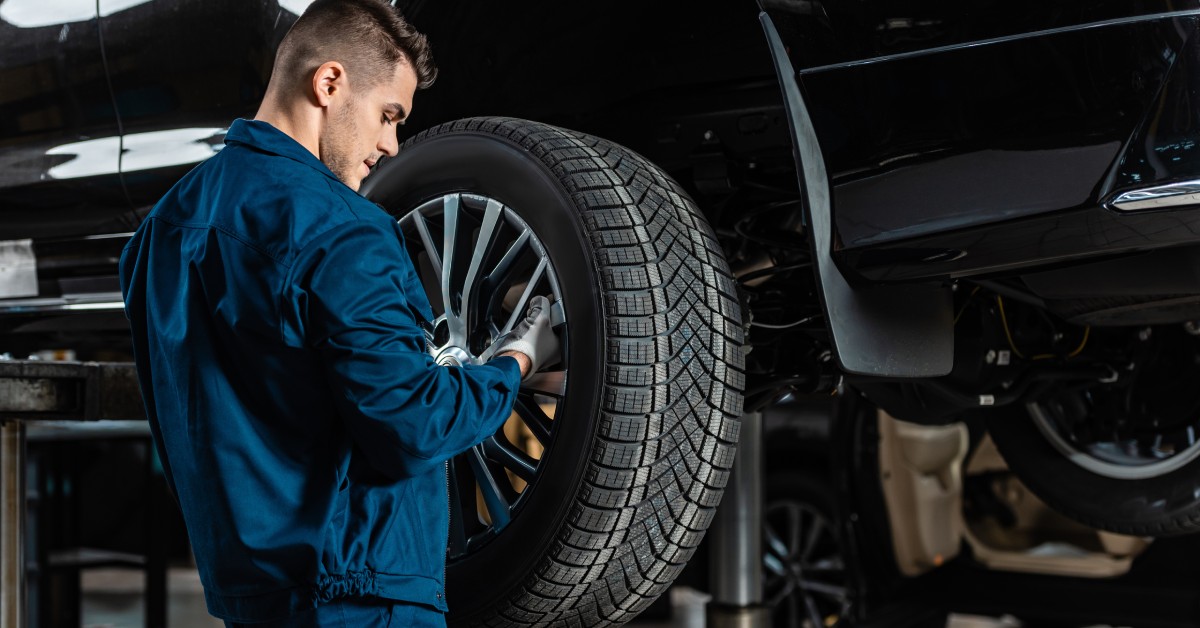A mechanic in an auto shop wearing a blue uniform with white gloves replacing a tire on a shiny black car.