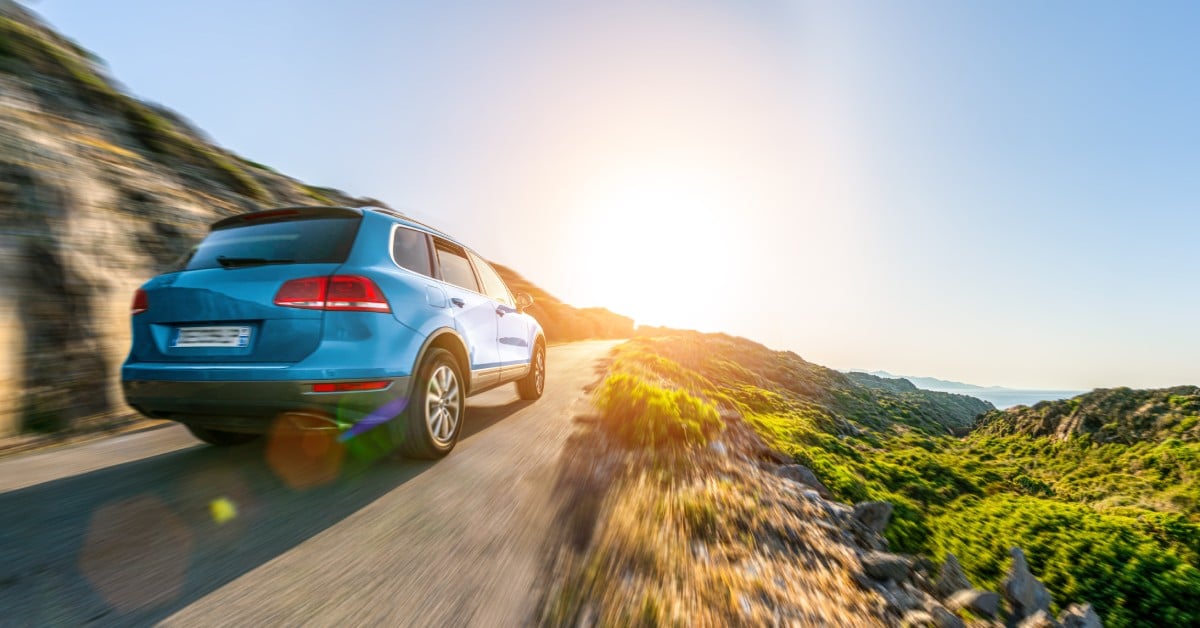 A blue SUV drives on a scenic coastal mountain road with motion blur and bright sunlight in the background.