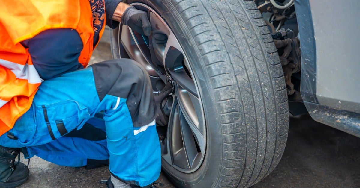 A mechanic in a blue and orange uniform performs tire rotation service on a vehicle wheel in a professional auto shop.