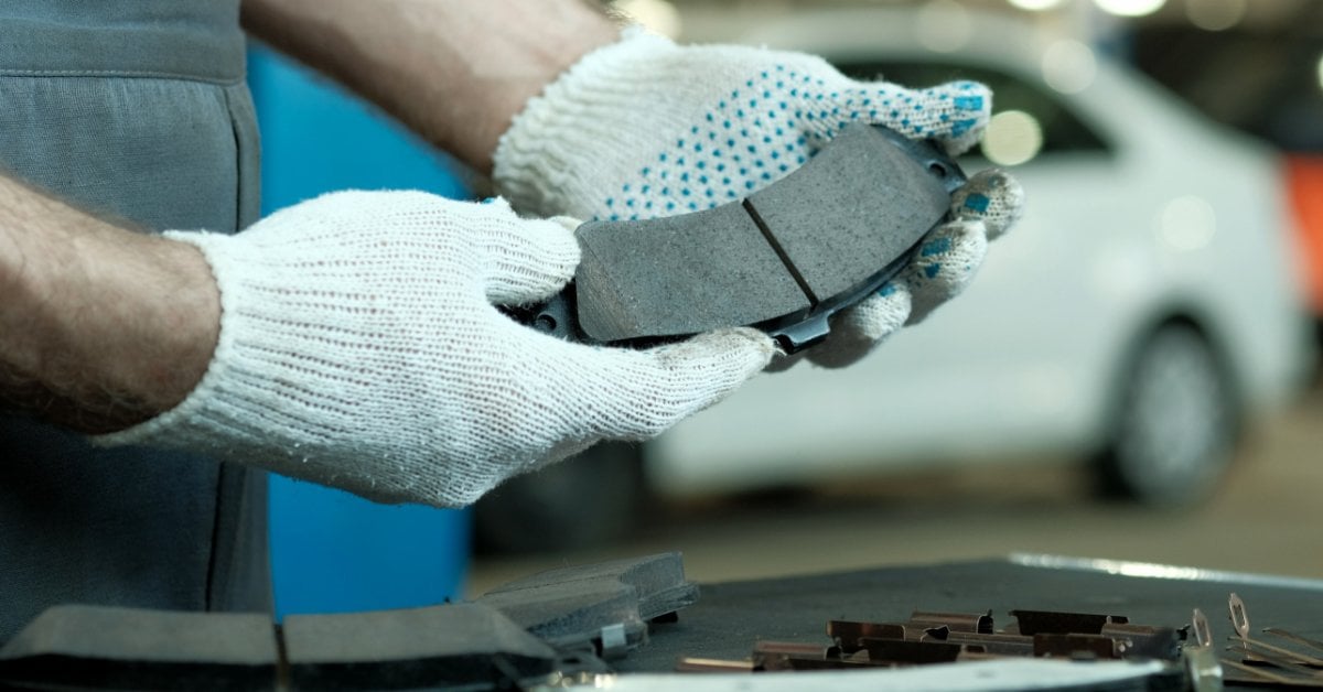 Semi-Metallic vs. Ceramic Brake Pads: Pros and Cons A mechanic wearing white gloves holds a new brake pad in an auto repair shop, with a white car blurred in the background.