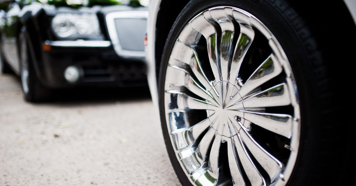 A close-up of a polished chrome wheel on a white vehicle with a black car blurred in the background.