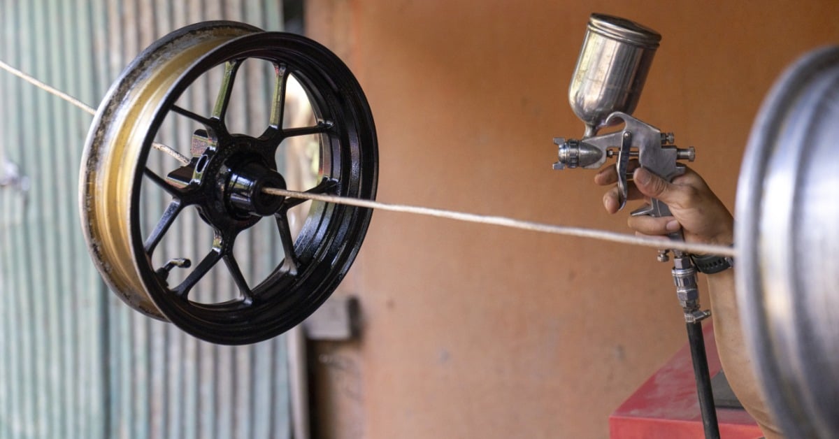 A technician uses a spray gun to apply a black painted finish to a vehicle wheel suspended by a wire.