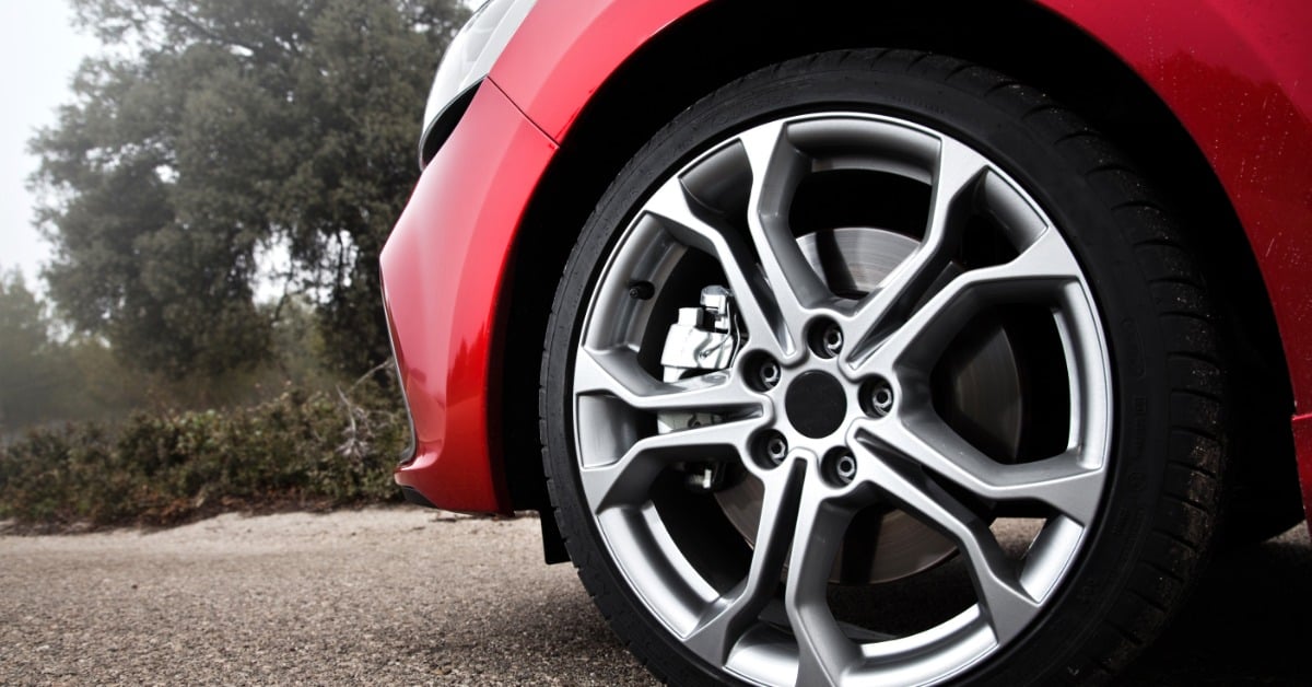 A low-angle view of a polished silver alloy wheel and tire on a red vehicle parked on a road with trees behind it.