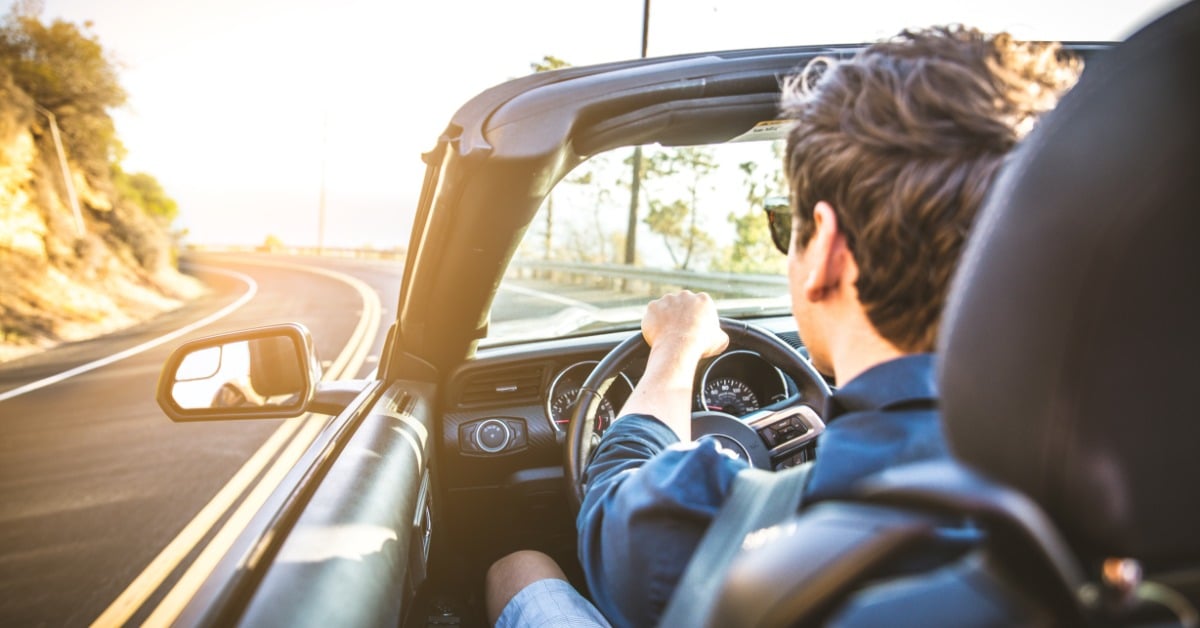 A man drives a convertible along a sunny winding road, gripping the steering wheel with bright sunlight flooding the scene.
