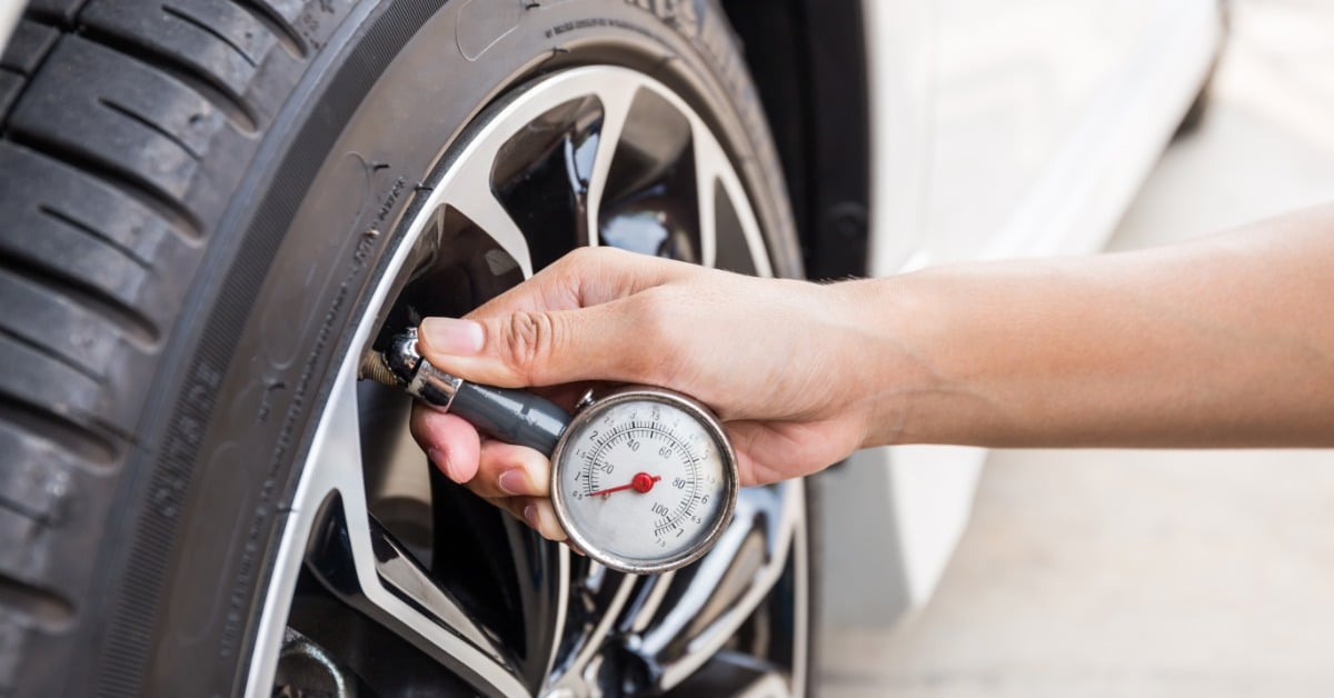 A hand holding a tire pressure gauge against a car tire valve stem, checking inflation levels on a white vehicle's alloy wheel.