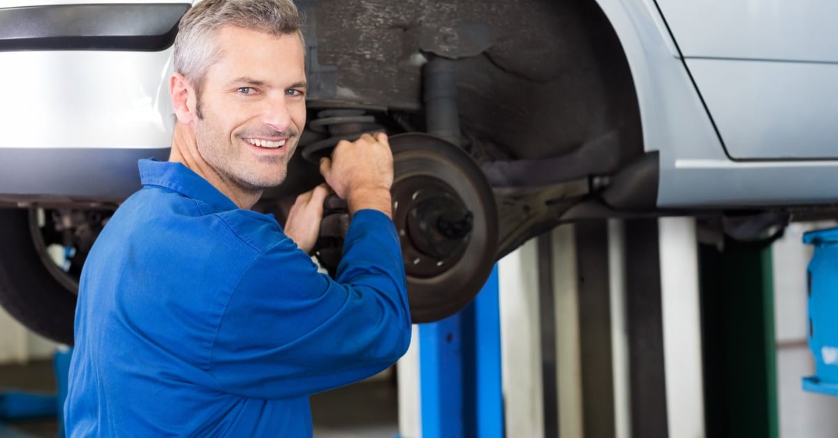 A mechanic in a blue uniform smiles at the camera while working on the brake rotor of a white car on a lift.