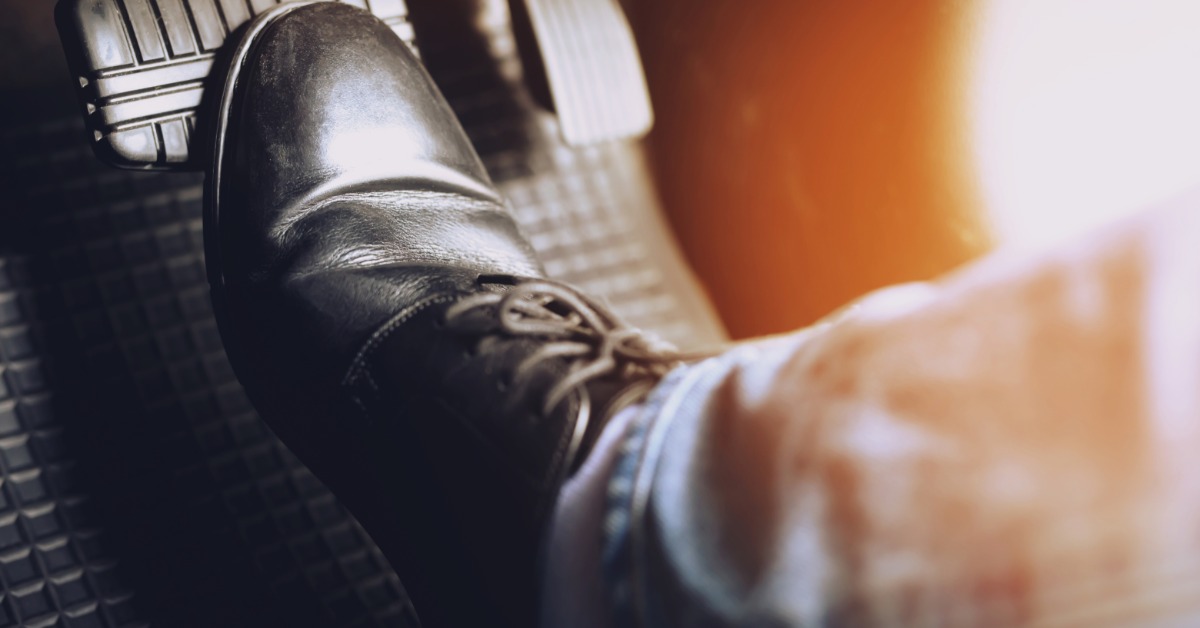 A close-up of a driver pressing a vehicle brake pedal with a black leather lace-up boot on a rubber floor mat.