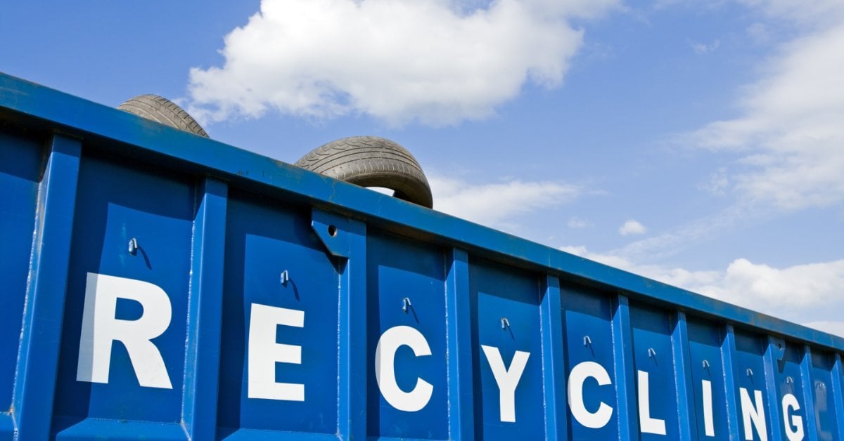 3 Tire Disposal Options Every Driver Should Know Old used car tires peek over the rim of a large blue recycling dumpster parked outside on a sunny, partly cloudy day.