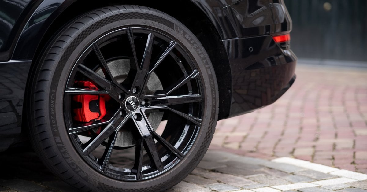 The Pros and Cons of Matte Black vs. Glossy Rims A close-up of a black alloy wheel with a red brake caliper on a car parked on cobblestone pavement.