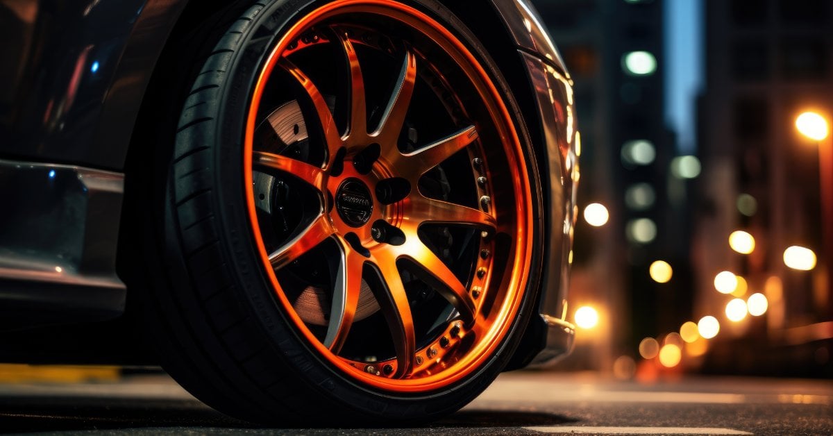 The Pros and Cons of Matte Black vs. Glossy Rims A close-up of an orange custom car wheel rim on a city street at night with blurred lights in the background.