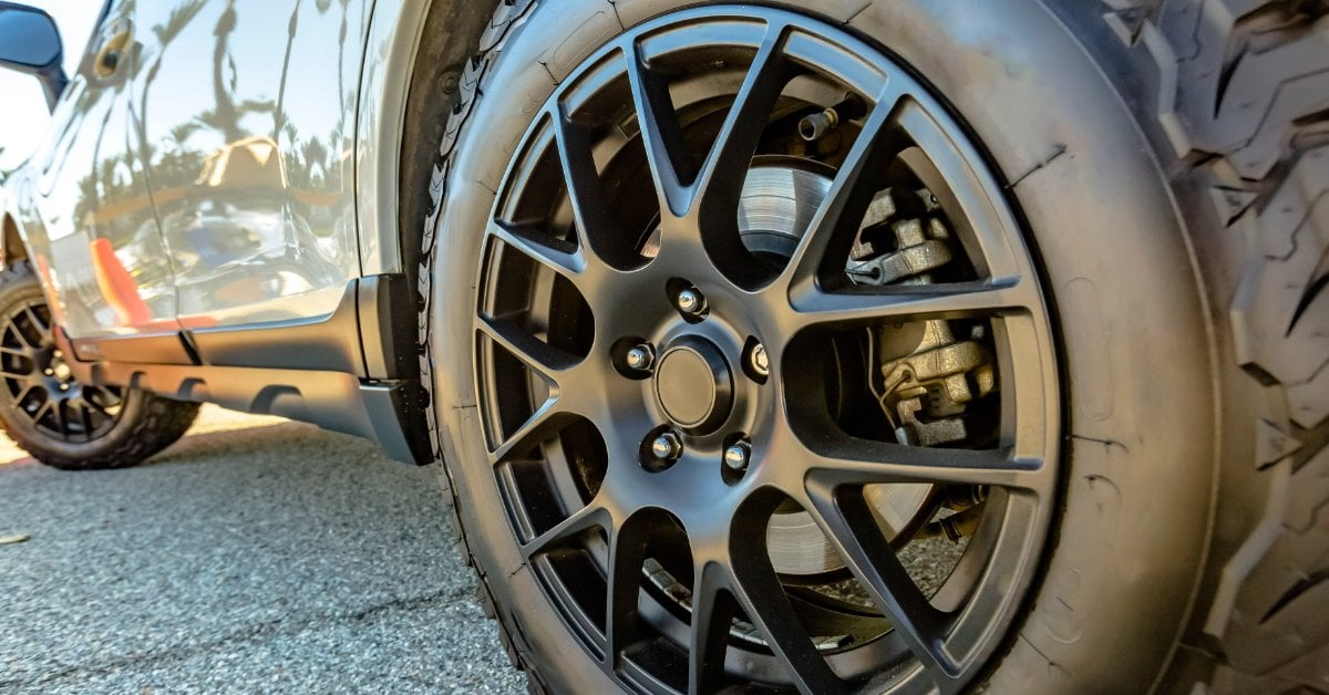 The Pros and Cons of Matte Black vs. Glossy Rims A close-up of a black alloy wheel with a rugged off-road tire on a parked SUV, with the brake caliper visible.